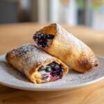 Two golden-brown air fryer blueberry chimichangas dusted with powdered sugar on a white plate.