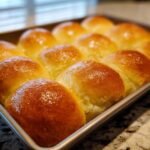 A close-up shot of a baking pan filled with golden-brown, fluffy dinner rolls, glistening with butter.