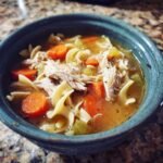 A close-up of a bowl of Cozy Crockpot Chicken Noodle Soup, featuring shredded chicken, wide egg noodles, carrots, and celery in broth.