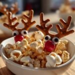 Close-up of a Cracker Jack Reindeer treat with pretzel antlers, candy eyes, and a red nose, nestled in popcorn.