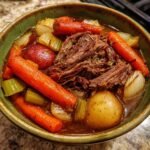 A hearty bowl of crock pot pot roast with tender shredded beef, carrots, potatoes, and onions in a rich broth.