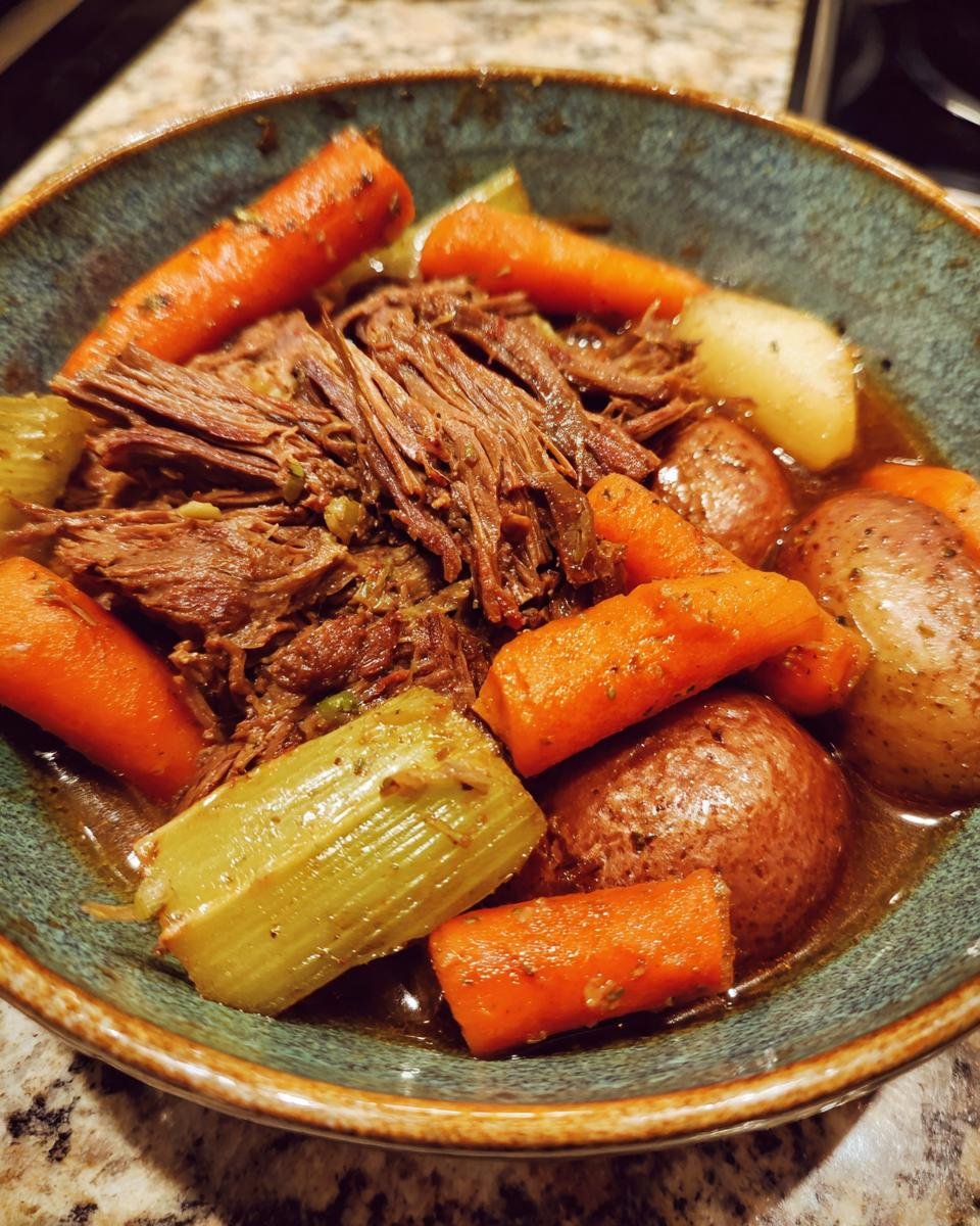 A close-up of a bowl filled with tender crock pot pot roast, carrots, celery, and potatoes in a rich gravy.