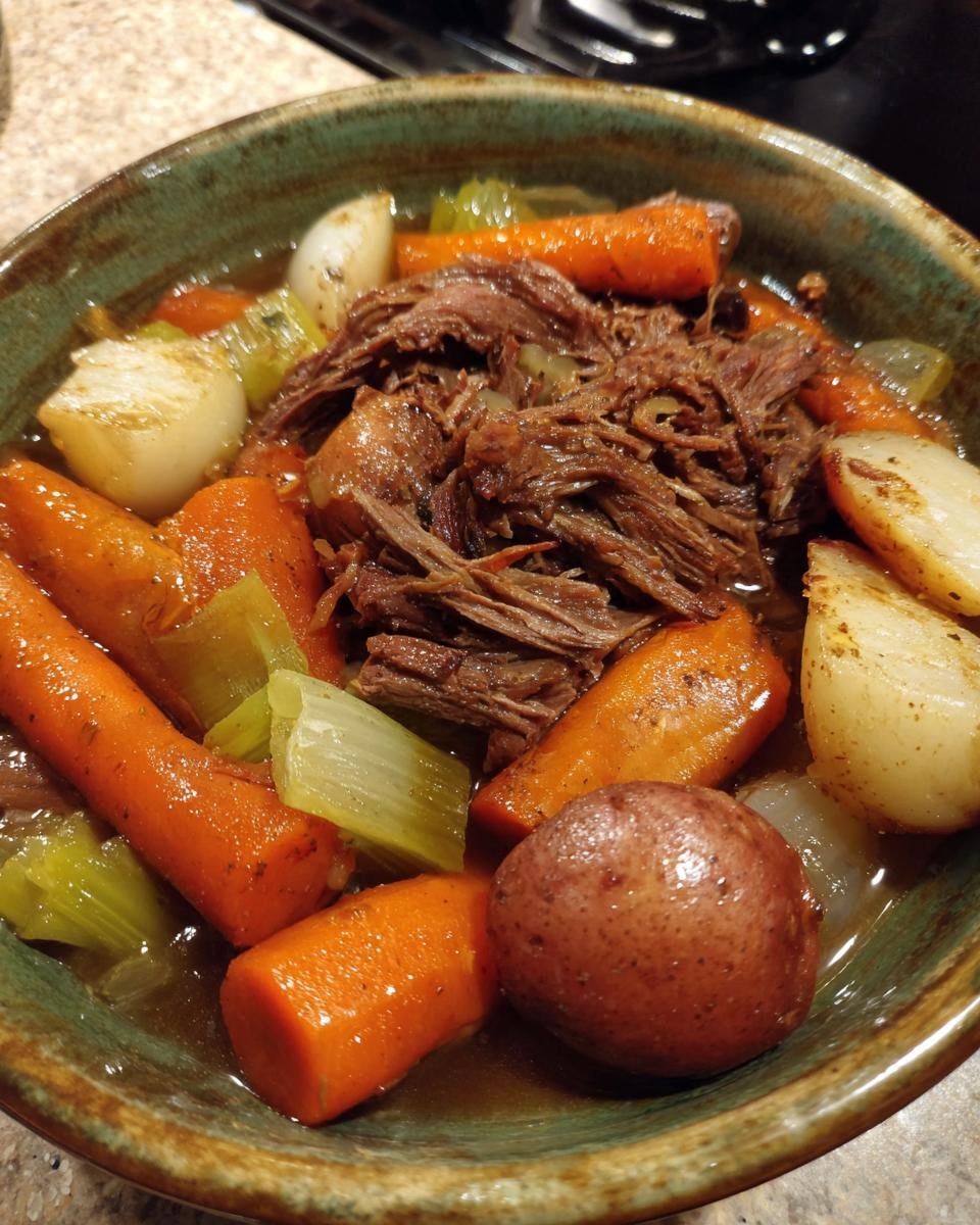 A close-up of a bowl filled with tender crock pot pot roast, carrots, potatoes, and celery in savory broth.