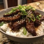A bowl of fluffy white rice topped with tender slices of glossy Easy Beef Teriyaki, garnished with sesame seeds and green onions.