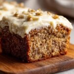 A slice of irresistible banana cake with cream cheese frosting and walnuts on a wooden board.