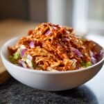 A close-up of an Irresistible Buffalo Chicken Salad Recipe in a white bowl, topped with shredded chicken and red onion.