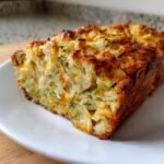A slice of irresistible savory zucchini cheddar quick bread on a white plate, showing visible zucchini shreds and melted cheddar cheese.