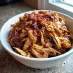 A white bowl filled with shredded Irresistible Slow Cooker Bacon Brown Sugar Garlic Chicken, topped with crispy bacon bits.