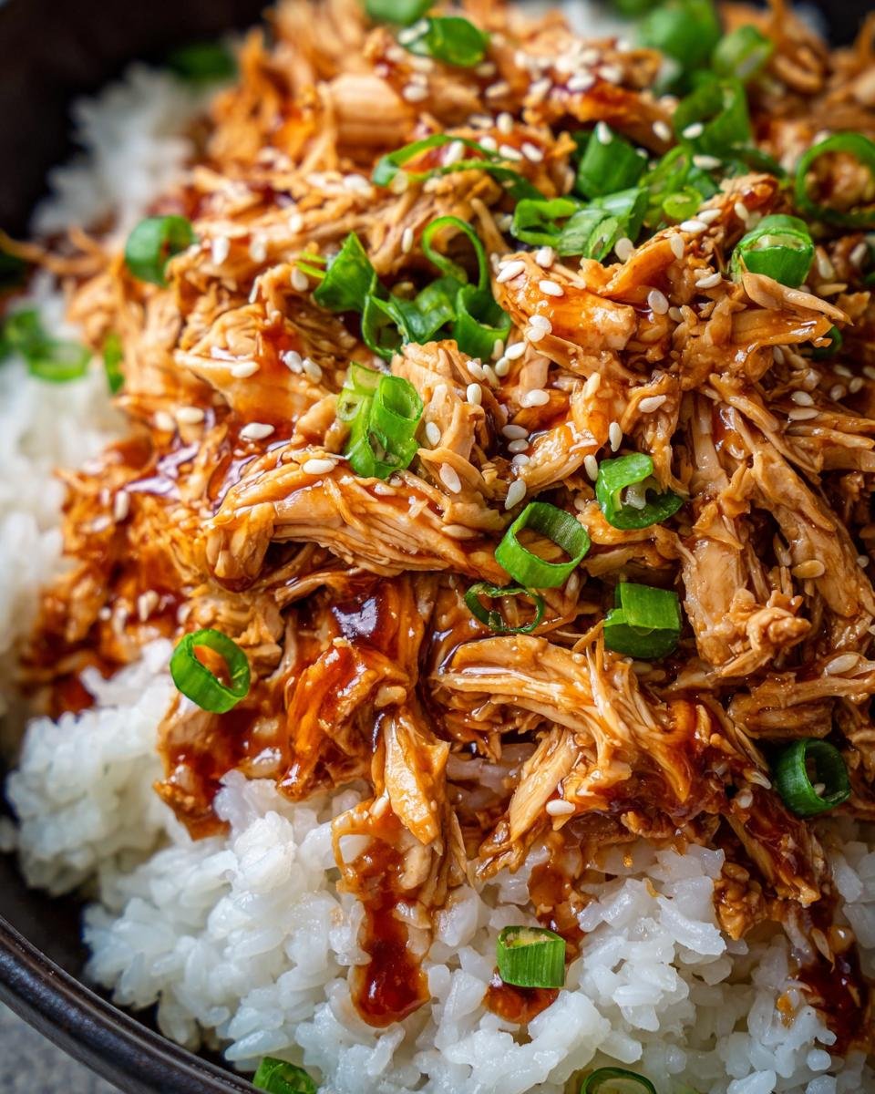 Close-up of shredded Irresistible Slow Cooker Honey Teriyaki Chicken served over white rice, garnished with sesame seeds and green onions.