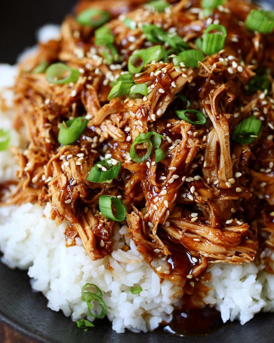 Close-up of shredded Irresistible Slow Cooker Honey Teriyaki Chicken served over white rice, garnished with sesame seeds and green onions.