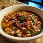 A close-up of a white bowl filled with Ultimate Bean with Bacon Soup, topped with crispy bacon and fresh herbs.