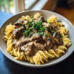A close-up of Ultimate Old Fashioned Beef Stroganoff Delight served over egg noodles, garnished with fresh parsley.