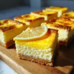 A close-up of a zesty lemon cream cheese bar, topped with a lemon slice, on a wooden board.