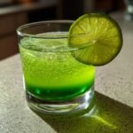 A vibrant green Alligator Cerveau cocktail in a glass, garnished with a lime slice and filled with tiny bubbles.