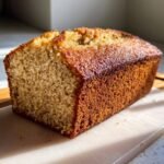 A golden-brown loaf of Brown Butter Bourbon Banana Cake, with a slightly cracked top, resting on a white cutting board.