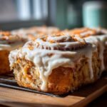 A close-up of a freshly baked Carrot Cake Cinnamon Roll generously topped with cream cheese frosting and shredded carrots.
