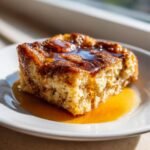 A golden square serving of Cinnamon Maple French Toast Bake drizzled heavily with maple syrup on a white plate.