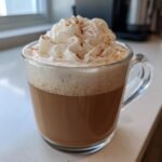 A warm Cinnamon Maple Vanilla Latte topped with whipped cream and a sprinkle of cinnamon in a clear glass mug.