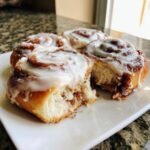 Close-up of fluffy Classic Homemade Cinnamon Rolls covered in thick white icing, one roll is broken open.
