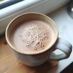 A close-up of a steaming mug of creamy almond butter hot chocolate with a frothy top.