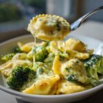 A fork lifting a piece of Creamy Broccoli Alfredo Ravioli, covered in rich cheese sauce and broccoli florets.