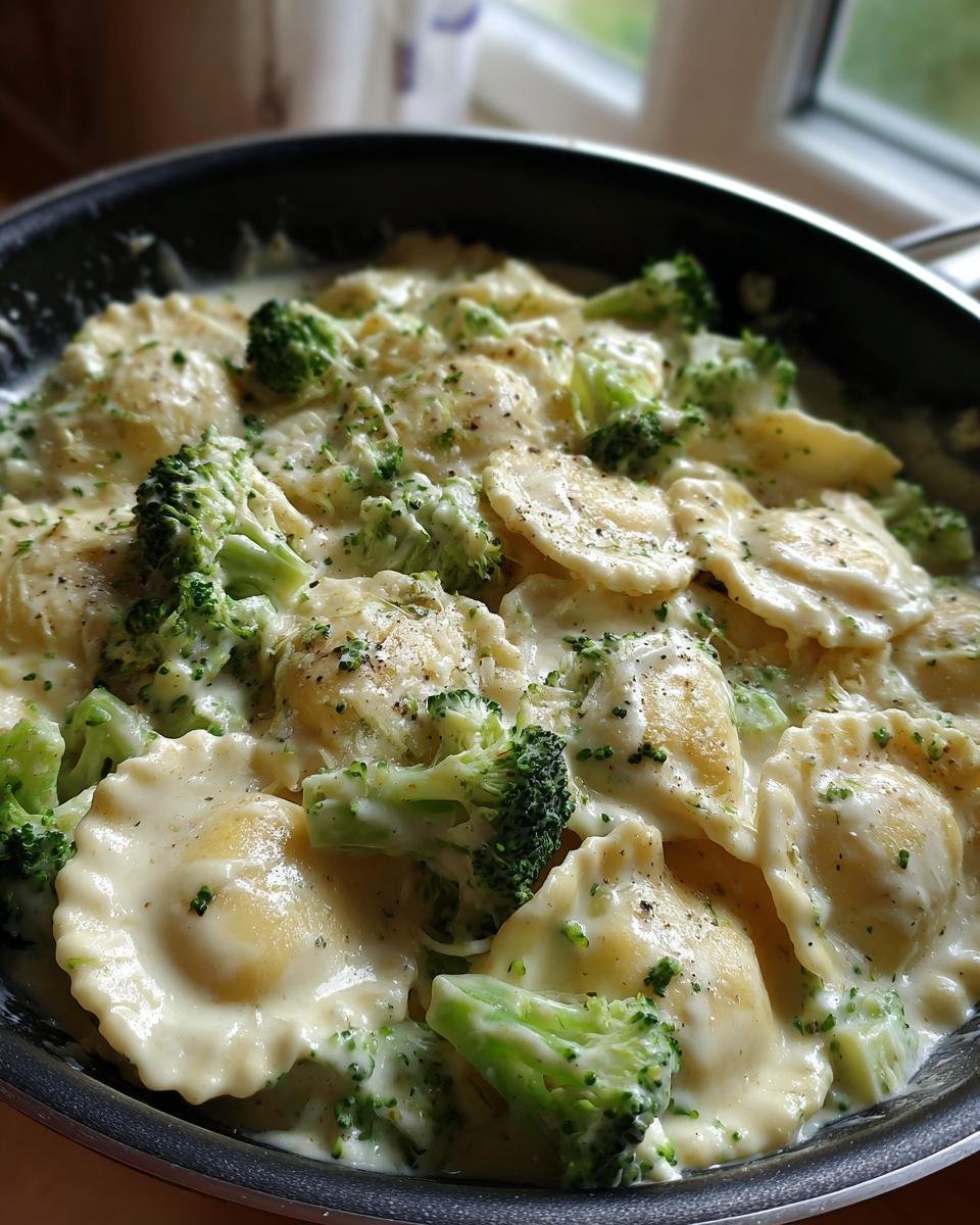 A close-up of Creamy Broccoli Alfredo Ravioli in a black pan, with vibrant green broccoli florets and a rich, creamy sauce.