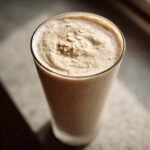 A tall glass filled with a creamy coconut oatmeal smoothie, topped with a sprinkle of oats.