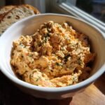 A close-up of a white bowl filled with creamy herb roasted salmon dip, garnished with dill. Slices of bread are in the background.