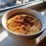 A close-up of a white bowl filled with creamy Jamaican cornmeal porridge, generously dusted with cinnamon.