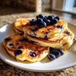 A stack of extra fluffy honey blueberry pancakes topped with fresh blueberries and syrup.