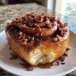 A close-up of a Giant Maple Bacon Cinnamon Roll, dripping with glaze and topped with crispy bacon bits, with a bite taken out.