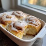 Close-up of freshly baked Banana Bread Cinnamon Rolls topped with thick white icing in a white baking dish.