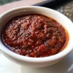 Close-up of a white bowl filled with vibrant, chunky homemade pizza sauce, showcasing herbs and spices.