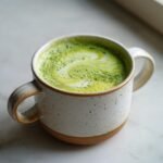A close-up of a frothy Honey Almond Matcha Latte in a speckled ceramic mug, showing swirls of green and white.