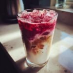 A tall glass filled with an Iced Strawberry Oat Milk Latte, showing layers of oat milk, coffee, and strawberry puree topped with ice.