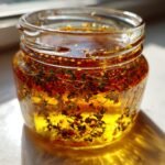 Close-up of bright yellow oil infused with herbs and red pepper flakes for Easy Homemade Italian Dressing in a small glass jar.