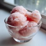 A clear glass bowl filled with scoops of pale pink lychee sorbet, with condensation on the bowl.