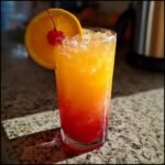 A tall glass filled with a vibrant Mango Orange Sunrise Mocktail, layered with orange and red hues, garnished with an orange slice and cherry.