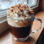 A clear glass mug filled with a Maple Pecan Sticky Bun Latte, topped with whipped cream and chopped pecans.