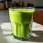 A vibrant green Matcha Pineapple Green Smoothie in a faceted glass, with foam on top.