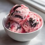 Three scoops of pink No Churn Strawberry Oreo Ice Cream studded with dark chocolate cookie pieces in a white bowl.