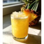 A tall glass filled with a bright yellow Pineapple Coconut Cream Mocktail, topped with crushed ice and a pineapple wedge garnish.