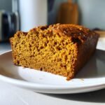 A close-up of a thick, moist slice of rich orange Pumpkin Chai Latte Bread on a white plate.