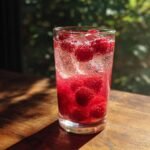 A tall glass filled with a vibrant Raspberry Lemonade Mocktail, packed with ice and fresh raspberries, sparkling with bubbles.