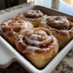 Four freshly baked Southern Peach Cobbler Cinnamon Rolls drizzled with white icing in a white baking dish.