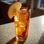 A tall glass filled with an amber Spiced Apple Bourbon Mocktail, ice, cinnamon sticks, and an apple slice garnish.