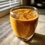 A close-up of a Spiced Apple Butter Oat Smoothie in a clear glass, with sunlight streaming in.