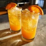 Two tall glasses filled with an orange Spiced Maple Citrus Pear Mocktail, ice, and garnished with fresh orange slices.