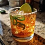 A refreshing spicy mezcal cocktail with jalapeno slices, lime wedge, and a salt rim, served over ice.