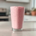 A tall glass filled with a thick, pink Strawberry Cheesecake Milkshake, showing condensation on the outside.